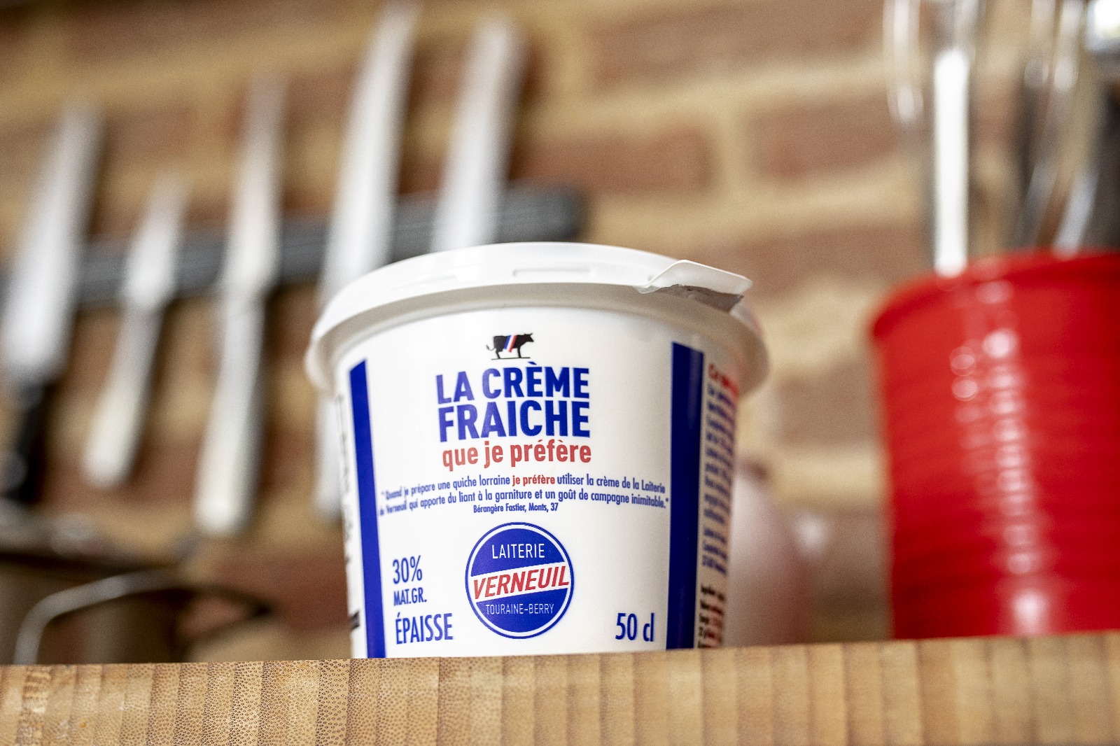 Pot de 50 cl de crème fraîche épaisse de la Laiterie de Verneuil posé sur un plan de travail en bois, en cuisine, avec des couteaux flous en arrière-plan.