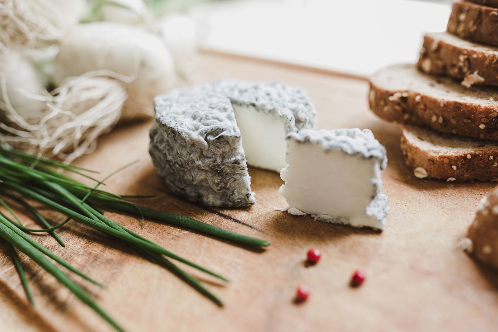 Fromage de chèvre cendré à croûte grise, partiellement coupé, posé sur une planche en bois avec de la ciboulette, du pain aux céréales et quelques baies roses.