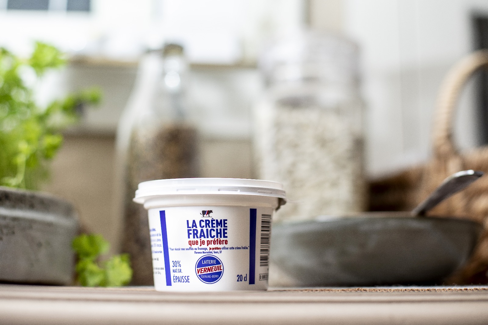 Pot de crème fraîche épaisse 20 cl de la Laiterie de Verneuil posé sur une table de cuisine, avec des herbes aromatiques et bocaux en arrière-plan.