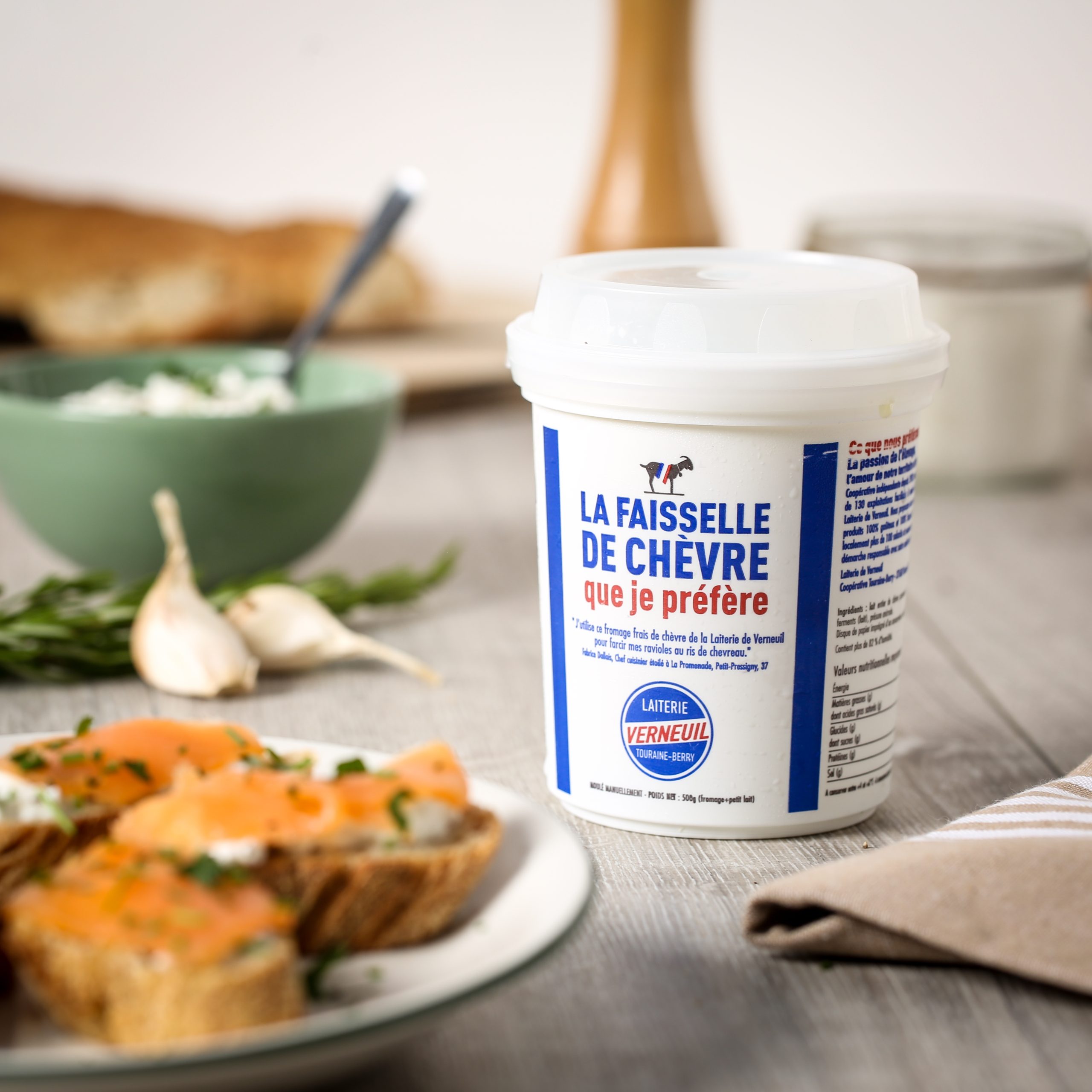 Pot de faisselle de chèvre de la Laiterie de Verneuil posé sur une table en bois, avec en arrière-plan de l’ail, des herbes, une tartine au saumon, et un bol de préparation à base de fromage frais.