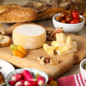 Plateau de fromage artisanal avec un fromage entier et des cubes coupés, entouré de fruits secs, pain, raisins, tomates cerise, figues et radis.