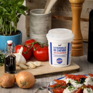 Pot de faisselle de chèvre de la Laiterie de Verneuil posé sur une planche en bois avec des tomates, ail, oignons, basilic et vinaigre balsamique.